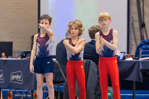 Three young gymnasts in matching uniforms watch intently, arms crossed and tense, awaiting results at a Gymfed event.