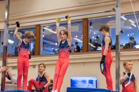 Three boys in red leotards hang and stand on gymnastics high bar, focused and determined, in a bright gymnasium.
