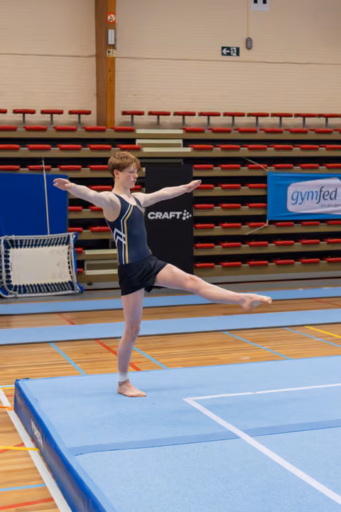 Young gymnast balances on one leg with arms extended, performing an arabesque on a blue floor mat in a sports hall.