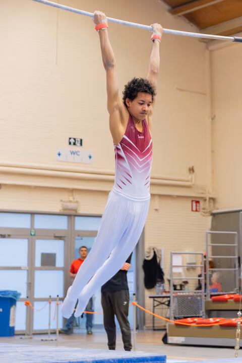 Young male gymnast hangs from the high bar with focused concentration, wearing a maroon and white leotard indoors.