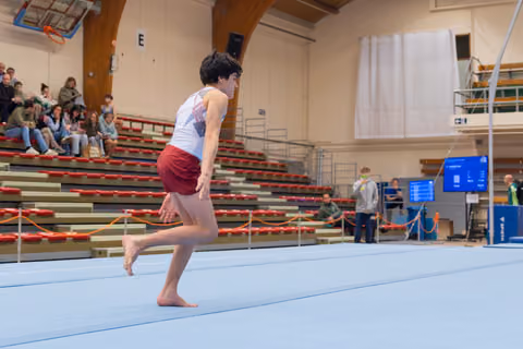Young male gymnast sprints barefoot across the blue floor mat, focused and poised, in a packed indoor gymnastics hall.