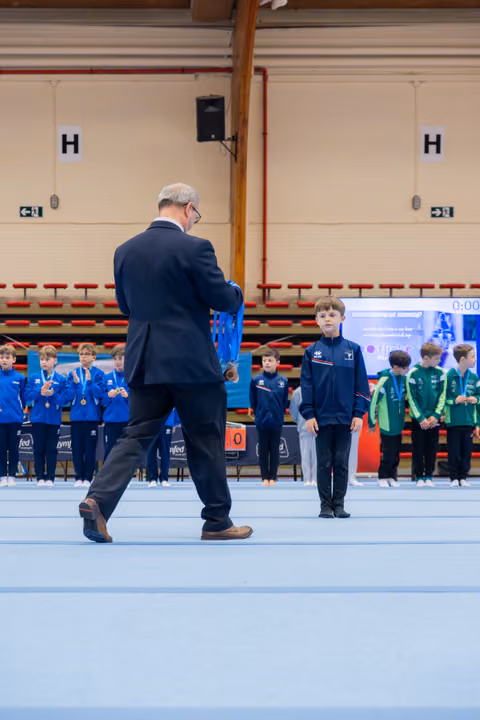An official in a suit approaches a young boy in a blue tracksuit to present a medal during a gymnastics awards ceremony.