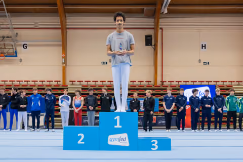 Young male gymnast stands proudly on the first-place podium, holding a trophy at a Gymfed indoor gymnastics event.