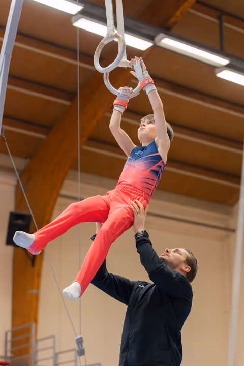 Young boy hangs focused on gymnastics rings while coach carefully spots him from below in an indoor gym.