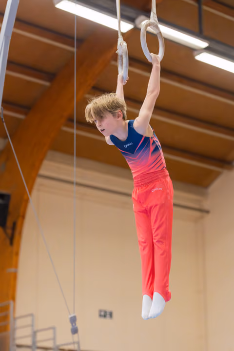 Young male gymnast hangs from still rings with focused expression, wearing red and blue Quatro leotard in a gymnasium.