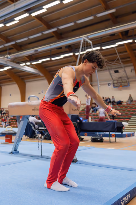 Young male gymnast landing on blue mat, leaning forward with arms extended, focused expression in indoor gymnastics hall.