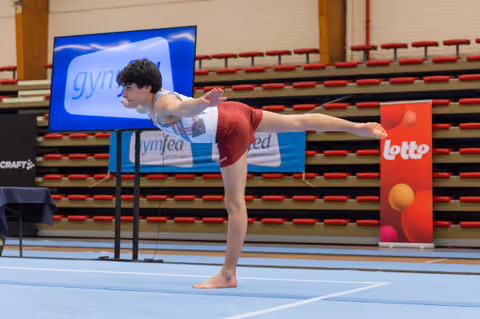 Young male gymnast holds a precise arabesque balance on the floor, arms extended, in a competitive gymnastics hall.