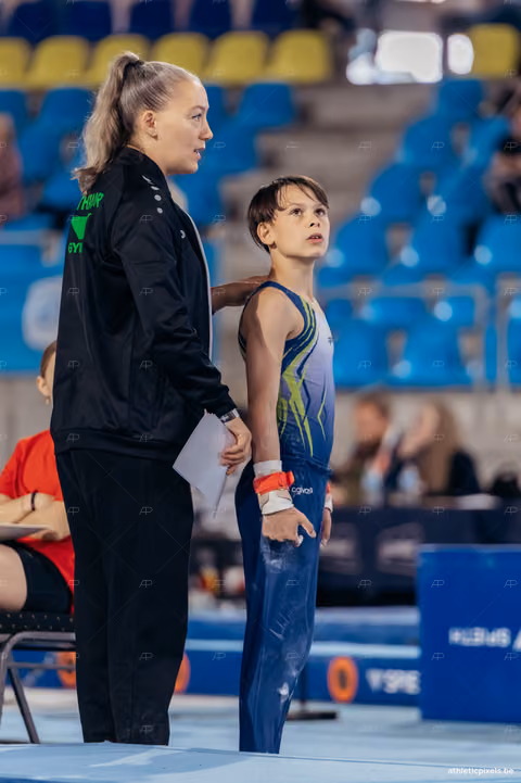 Young gymnast stands focused on the floor, coach placing a reassuring hand on their back before their routine.
