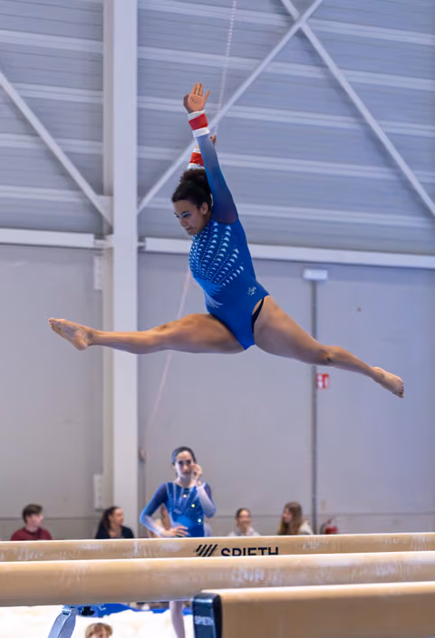 Gymnast executes a split leap with arms extended on balance beam during indoor training, showcasing flexibility and form