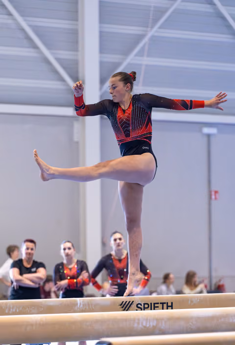 Athlete performing on balance beam with extended leg and arms, teammates in matching outfits watching in background