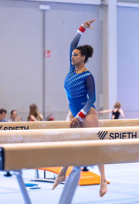 Female gymnast performing on balance beam with one arm raised, wearing blue leotard with red and white accents