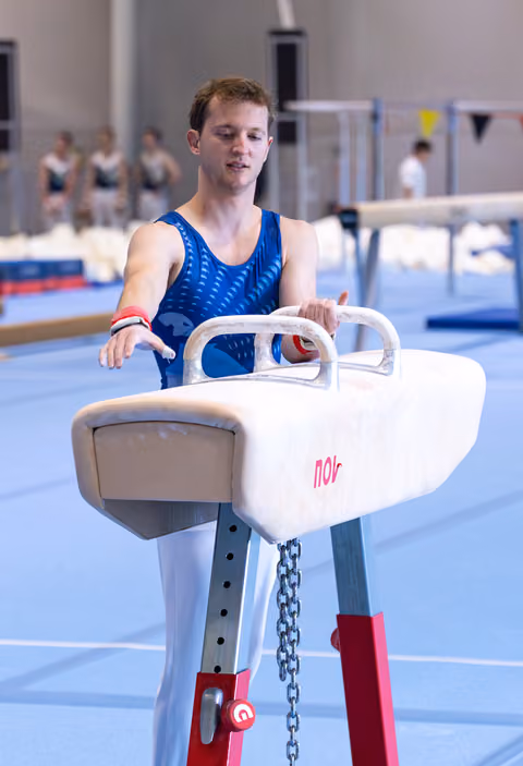 Young male gymnast grips pommel horse handles during training, focused expression, wearing blue leotard in indoor facility
