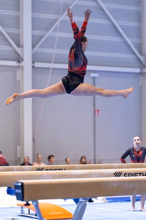 Gymnast performs a perfect split leap over the balance beam with arms raised, showcasing strength and flexibility.