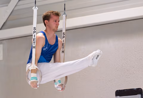 Young male gymnast performing a horizontal leg position on still rings, wearing blue leotard and white pants