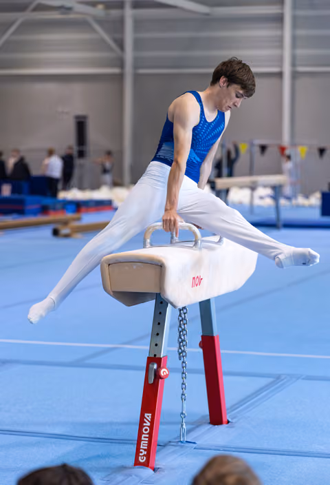 Young male gymnast performing a split position on pommel horse, demonstrating strength and control in training facility