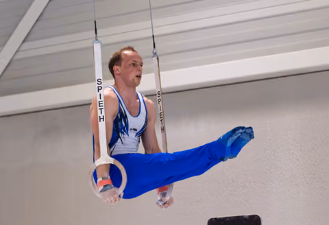 Male gymnast executing a horizontal hold on still rings, legs extended in blue attire, displaying intense concentration