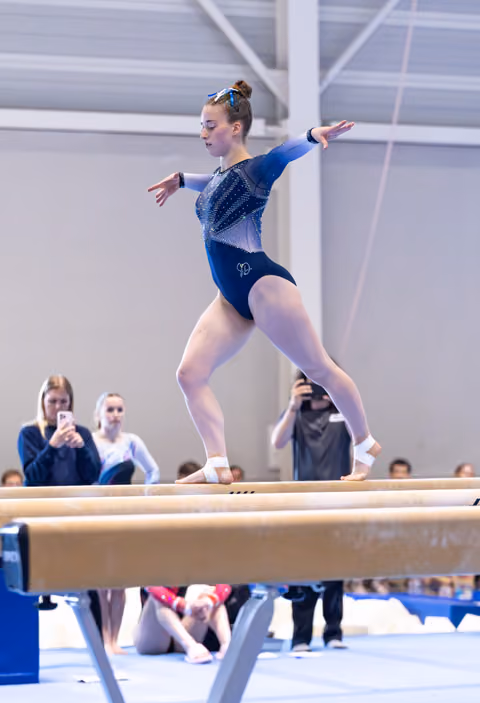 Gymnast executes artistic pose on balance beam with arms extended, wearing blue leotard while spectators watch