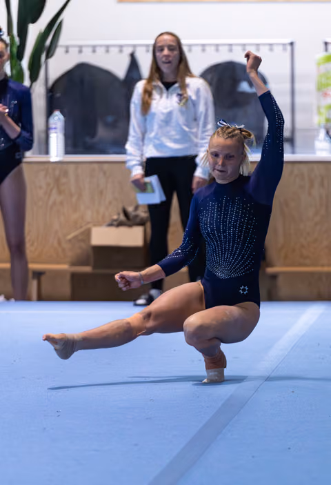 Young gymnast performing a floor routine landing pose in navy leotard while coach observes in background