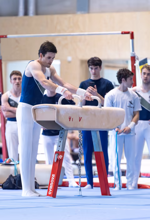 Athlete performs pommel horse routine with focused concentration while teammates watch in background at training facility