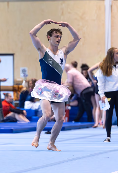 Young man performs expressive dance routine with heart-shaped hand gesture above head, wearing colorful tutu on floor exercise mat