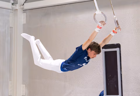 Young athlete demonstrates impressive horizontal hold on still rings, arms extended overhead, wearing blue uniform and white pants