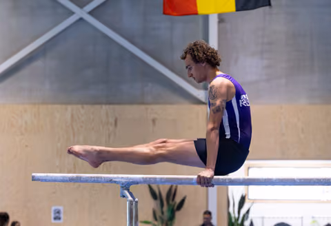 Male athlete performing L-sit on parallel bars, displaying strength and control in purple sleeveless uniform