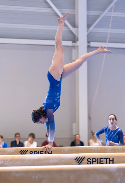 Gymnast performing inverted handstand with vertical leg splits on balance beam during routine at indoor facility
