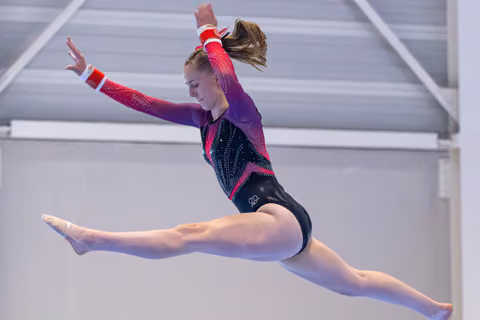 Gymnast performs a soaring split leap with extended arms, wearing a purple and black leotard in mid-air
