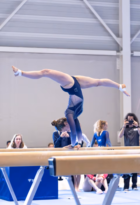 Young athlete performs a one-armed handstand on balance beam, legs split gracefully, while spectators watch in indoor venue