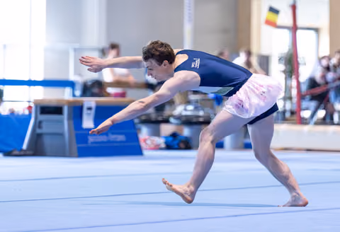 Male athlete performs dynamic leap during floor routine, arms extended with intense concentration at indoor sports facility