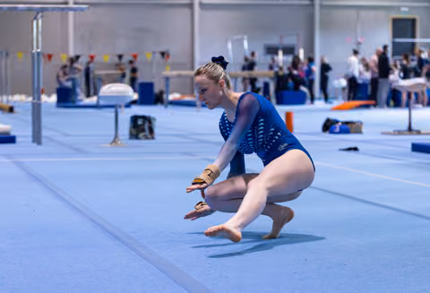 Young athlete executing a floor routine landing on blue mat, arms extended mid-performance in training facility