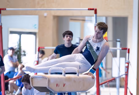 Male athlete performing pommel horse routine with extended legs while coach observes in indoor training facility