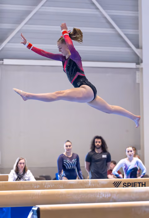 Young athlete performs a dynamic split leap above balance beam during training, with coaches and teammates observing below