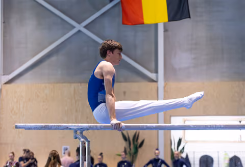 Athlete performing horizontal balance on parallel bars with German flag visible in background during training session