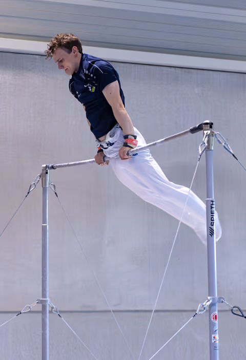 Male athlete performs on horizontal bar, body extended horizontally in white pants and navy shirt against industrial wall
