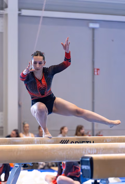 Gymnast performs focused routine on balance beam with extended leg and raised arms, displaying concentration and control