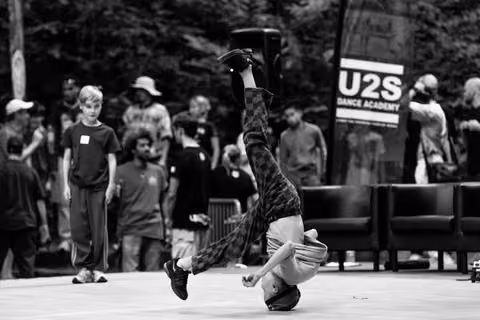 Breakdancer executes an inverted headstand move on stage, with audience and U2S Dance Academy banner visible in background