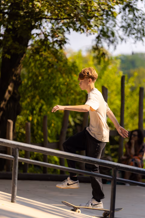 Young skater balances on rail during grind trick at outdoor skatepark, focused expression, surrounded by lush greenery