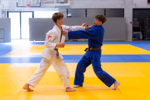 Two young judoka practice gripping techniques on yellow training mats in a modern sports facility, wearing white and blue gi
