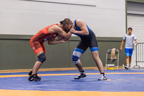 Two male wrestlers in intense clinch, red vs blue singlet, both leaning in aggressively on an orange and blue mat.