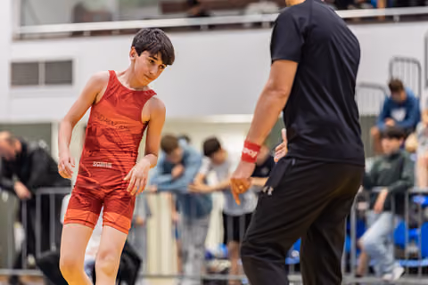 Young wrestler in red singlet crouches in focused stance, facing an adult coach on the mat during a sports event.