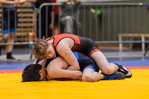 Teenage wrestler in red singlet pins dark-haired opponent to yellow mat, applying controlled pressure during match.