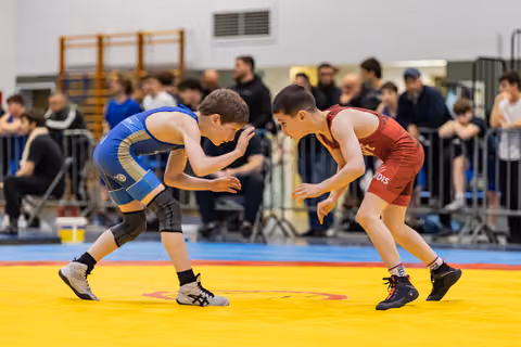 Two boys in blue and red singlets crouch in focused stance, circling each other on a yellow mat before a crowd.