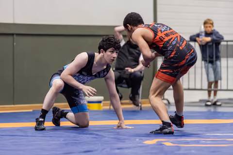 Teen wrestler in blue singlet crouches defensively on the mat, facing opponent in black-red uniform, both intense and focused.