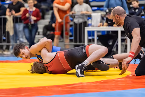 Young wrestler in red singlet pins opponent to colorful mat as referee leans in closely to observe the hold.