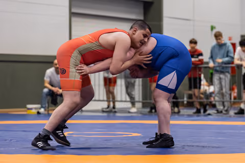 Two young wrestlers lock arms in an intense clinch on a blue mat, straining forward with determined effort.