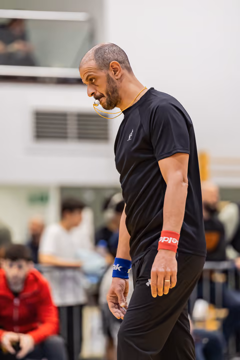 Male referee with whistle in mouth leans forward intently, wearing black shirt and red wristband in indoor sports hall.
