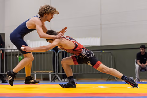 Curly-haired wrestler in blue singlet defensively pushes back opponent lunging low in red, on colorful mat.