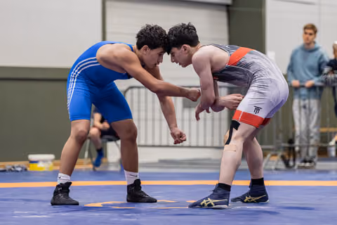 Two young wrestlers in blue and gray singlets lock up forehead-to-forehead in an intense grappling standoff on a blue mat.