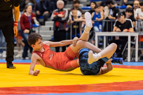 Boy in red singlet grips opponent's leg during intense floor grapple on colorful competition mat, crowd watching.